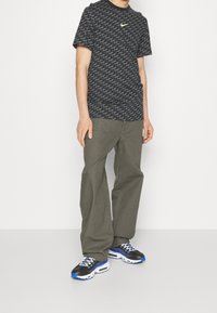 Černé grafické tričko s bílým a šedým vzorem vlny, logo v zeleném akcentu; olivově zelené volné kalhoty; černé a modré tenisky.