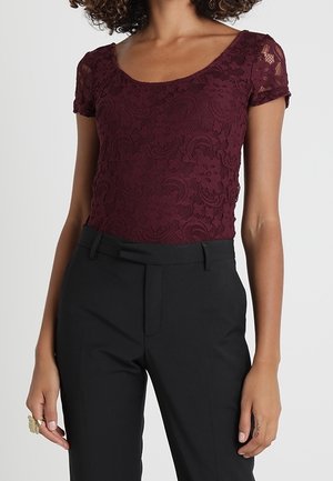 Kvinde iført en kortærmet bordeaux blondebluse med rund hals, stoppet ned i højttaljede sorte skræddersyede bukser.