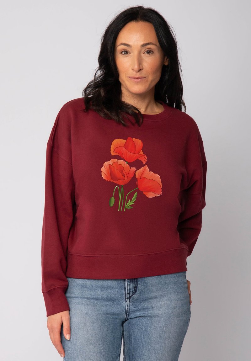 Vrouw met donker haar, gekleed in een bordeauxrode sweatshirt met rode klaprozen bedrukt op de voorkant en een lichtblauwe spijkerbroek.