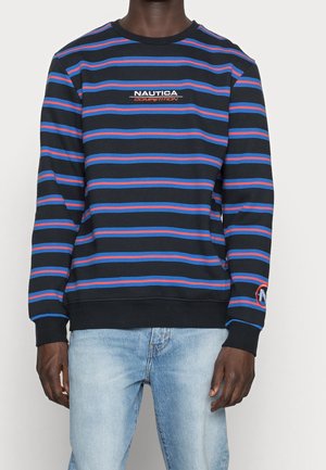 Mand iført sort crewneck sweater med blå og røde vandrette striber og "Nautica Competition" logo, kombineret med lyseblå jeans.