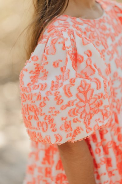 Robe brodée pêche avec des manches courtes bouffantes, présentant des motifs floraux intricats sur un tissu léger et transparent.
