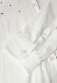 Witte shirt met een frisse textuur, voorzien van zilveren studs op de borst en een manchet met een heldere knoopdetail. Glad stofafwerking.