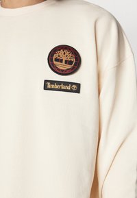 Sudadera crema con cuello de canalé, que presenta un parche redondo negro y rojo con un logo de árbol y un parche rectangular negro con "Timberland" en dorado.