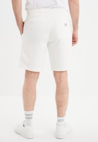 Witte katoenen shorts met een elastische tailleband, omslaghem, en een enkele achterzak met een klein logo-label. Draagt met witte sneakers.