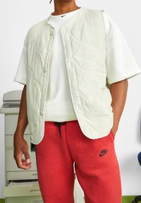 Ljust vadderad väst i off-white med rund halsringning och tryckknappar, bärs över en vit t-shirt och röda sweatpants, med Nike-logotyp.