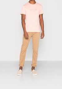 Camiseta de algodón rosa claro con el logo "HILFIGER", combinada con pantalones chinos beige y zapatos slip-on ligeros. Diseño minimalista, ajuste casual.