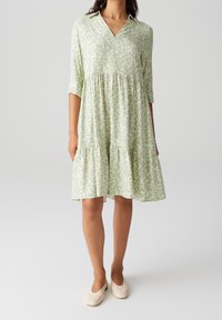 Robe verte à motifs avec un col en V, des manches trois-quarts et un ourlet à volants. Le tissu semble léger avec une texture douce.