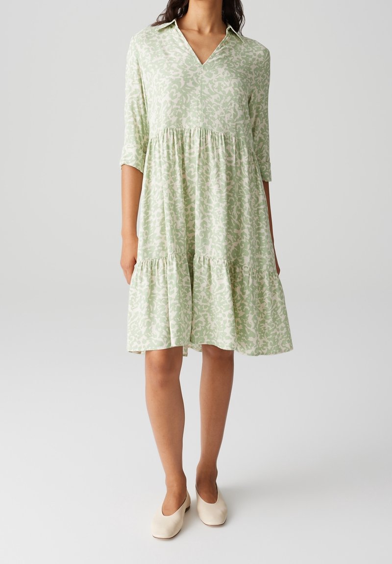 Robe verte à motifs avec un col en V, des manches trois-quarts et un ourlet à volants. Le tissu semble léger avec une texture douce.