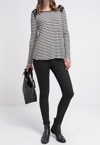 Svart och vit randig långärmad topp med spetsdetaljer på axlarna, parat med svarta skinny jeans och ankelstövlar. Håller en svart handväska.