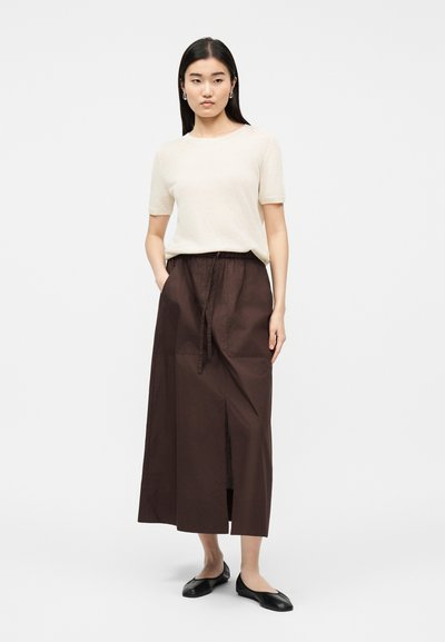 Femme debout, mains dans les poches, portant un haut crème à manches courtes, une longue jupe marron foncé fendue sur le devant, et des ballerines noires.