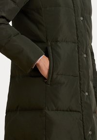 Tumši zaļš pūkains mētelis ar quilted dizainu, kurā ir roka sānu kabatā un snap-button aizdare. Gluda un gluda tekstūra.