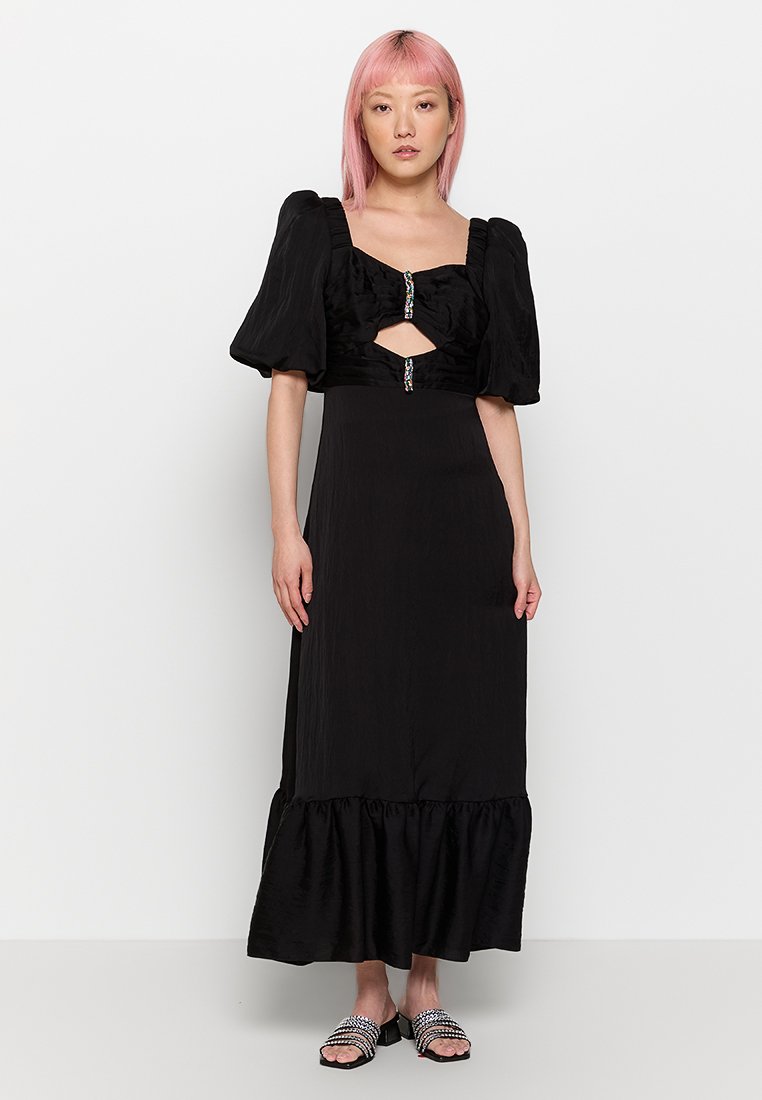 Frau mit pinken Haaren, die ein schwarzes Maxikleid mit Puffärmeln, Ausschnitten und mit Schmuckknöpfen trägt, kombiniert mit schwarzen, verzierten Sandalen mit offenen Zehen.