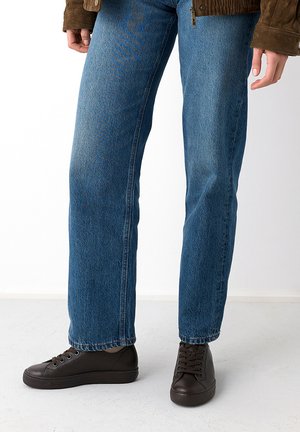 Person trägt blaue Jeans mit geradem Bein, braune Sneaker und eine braune Jacke und steht auf einem hellen Boden vor weißem Hintergrund.