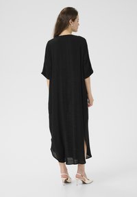 Zwarte, mid-length jurk met korte mouwen, zijspleten en een relaxte pasvorm. De stof lijkt lichtgewicht met een gestructureerde afwerking. Draagt bij beige hakken.
