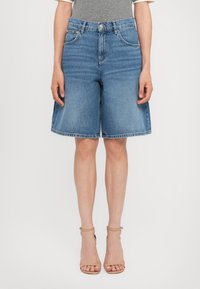 Pantalones cortos de mezclilla en azul claro, con una cintura alta, pasadores para cinturón, bolsillos frontales y un dobladillo sin terminar. Combinados con sandalias de tiras al tobillo en color beige.