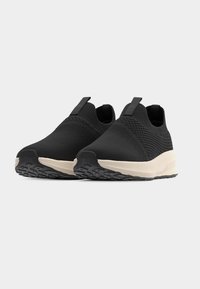 Ζευγάρι μαύρα αθλητικά παπούτσια  χωρίς κορδόνια με υφασμάτινα επάνω μέρη με υφή και λευκές ενισχυμένες σόλες σε απλό φωτεινό φόντο.