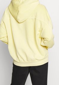 Žlutá mikina s kapucí na šňůrku, spadlými rameny, žebrovanými manžetami a lemem. Tkanina vypadá měkká a texturovaná, navržena pro pohodlí.