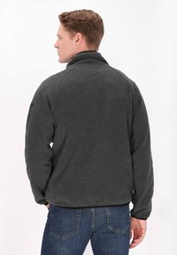 Giacca in pile grigio scuro con colletto alto, maniche raglan e finitura testurizzata. Presenta un orlo nero ed è abbinata a jeans blu.