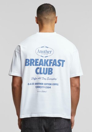 Hvit bomull T-skjorte med blå tekst som leser "Another Cotton Lab" og "Breakfast Club" på baksiden, med ekstra kontaktopplysninger nedenfor.