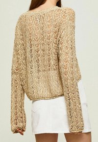 Beige gehäkelter Pullover mit langen Ärmeln und lockerer Passform, mit einem strukturierten Muster und einem leicht verkürzten Rücken. Kombiniert mit einem weißen Rock.