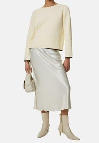Kremowy sweter o teksturze z prążkowanymi wykończeniami, połączony z satynową beżową spódnicą midi. Modelka trzyma lekką torebkę i nosi beżowe botki za kostkę.