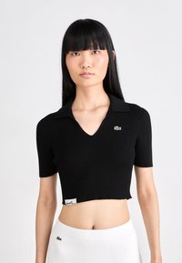 Top scurt cu mânecă scurtă, dintr-un material ribbed, de culoare neagră, cu decolteu în V, având un mic logo verde pe piept și o etichetă la tiv.
