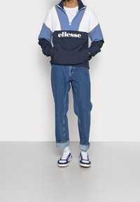 Marineblauw en lichtblauw jack met halve rits en wit logo, gecombineerd met opgerolde blauwe spijkerbroek en witte sneakers met donkere accenten.