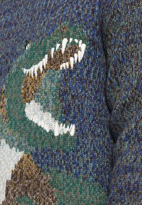 Pulover tricotat cu un design reprezentând un dinozaur verde și maro, având dinți albi, plasat pe un fundal texturat în nuanțe multicolore de albastru și maro.