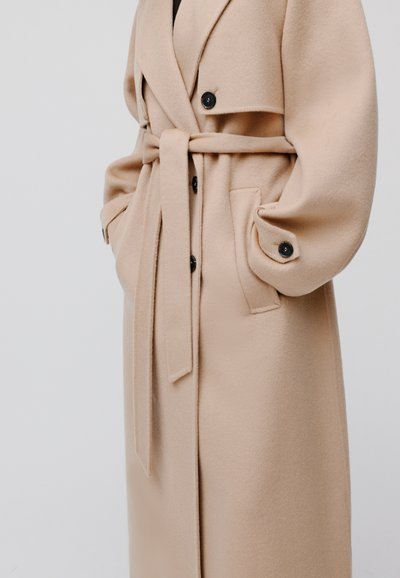Cappotto in misto lana beige con design doppiopetto, grandi bottoni neri, vita con cintura e tasche laterali. Tessuto liscio e maniche oversize.