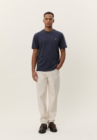 Tamsiai mėlynas trumpomis rankovėmis marškinėliai su apvalia iškirpte ir subtiliu logotipo detale ant krūtinės, derinami su šviesios spalvos kelnėmis ir rudais batais.