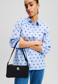 Sac bandoulière noir avec fermeture éclair et accent de personnage de dessin animé, associé à une chemise rayée bleu clair avec de petits motifs noirs.