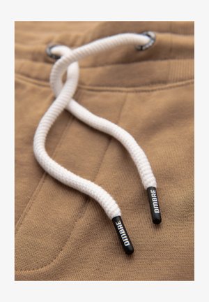 Tessuto marrone con una superficie texture presenta corde di chiusura bianche. Le estremità delle corde di chiusura sono in plastica nera con il testo "DIDAMBRE".