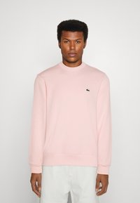 Sweatshirt rose clair en tissu doux, avec un col rond côtelé et des manches longues. Comprend un petit logo noir sur la poitrine.