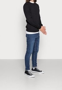 Un pull en maille noir superposé à une chemise blanche, associé à un jean slim-fit bleu et des baskets montantes noires avec des semelles blanches.