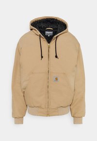 Beżowa bluza rozpinana z czarną pikowaną podszewką, regulowanym kapturem ze sznurkiem, dużymi przednimi kieszeniami, ściągaczami na mankietach oraz naszywką z logo po lewej stronie.