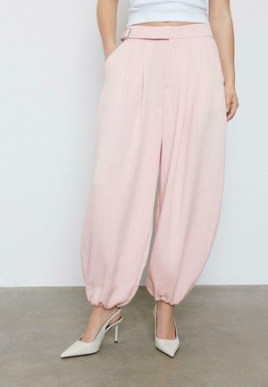 Žena stojaca na šedom povrchu, oblečená v svetloružových nohaviciach s vysokým pásom a širokými nohavicami so stiahnutými lemy, a bielych lodičkách na päte s špicatou špičkou.