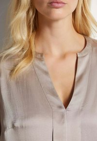 Beige zijden blouse met V-hals en korte mouwen. Voorzien van subtiele textuur en stiksels langs de halslijn en voorkant.