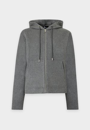 Un mannequin masculin se tient devant un fond blanc uni, vêtu d’un sweat à capuche gris zippé, d’un tee-shirt blanc, d’un jean gris ample et de chaussures noires.