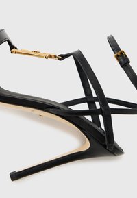 Μαύρο σανδάλι με ψηλό τακούνι, με πολλαπλά διασταυρούμενα λουριά, λείο δερμάτινο υφή και μεταλλικό διακοσμητικό σε χρυσό τόνο με κούμπωμα.