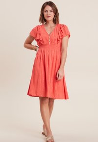 Korallenfarbenes Kleid mit Puffärmeln, V-Ausschnitt und elastischem Bund. Verfügt über subtile metallische Akzente und hat eine Länge bis zum Knie. Angenehme, glatte Textur.