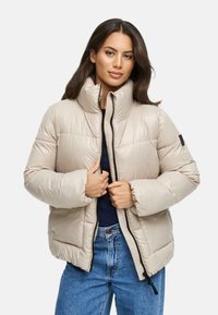 Beige pufferjack met een hoge kraag, zwarte ritssluiting en elastische manchetten; gecombineerd met denim jeans met een gladde, glanzende textuur.