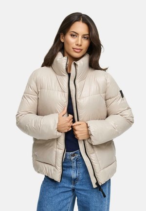 Beige pufferjack met een hoge kraag, zwarte ritssluiting en elastische manchetten; gecombineerd met denim jeans met een gladde, glanzende textuur.