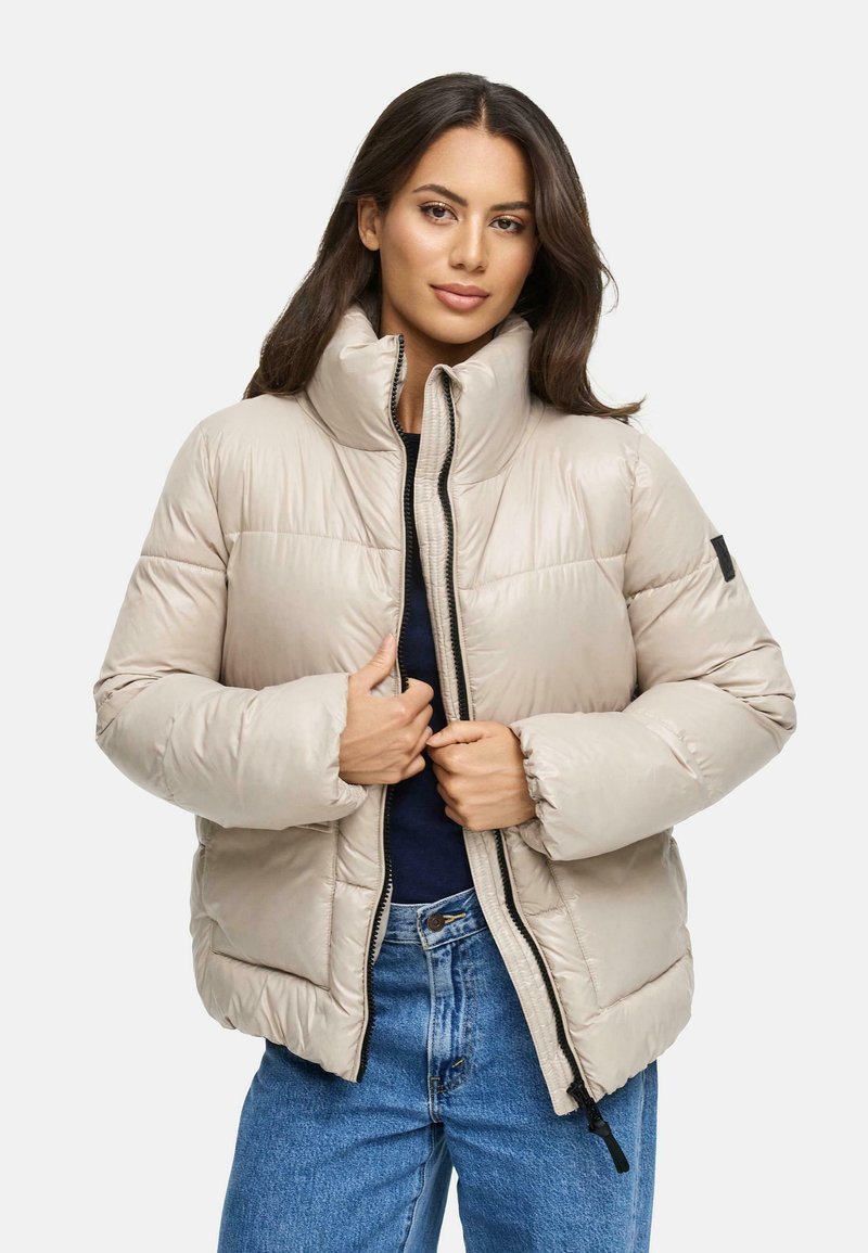 Beige pufferjakke med høj krave, sort lynlås og elastiske manchetter; parret med denimjeans, der har en glat, skinnende tekstur.