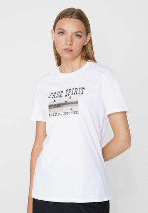 Jeune femme aux cheveux raides portant un t-shirt blanc avec des oiseaux et le texte "FREE SPIRIT NO RULES, JUST VIBES" sur un fond clair.