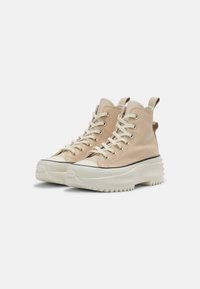 Beige leren sneakers met hoge schacht, dikke witte plateauzolen, beige veters, metalen oogjes en trekriemen op een witte achtergrond.