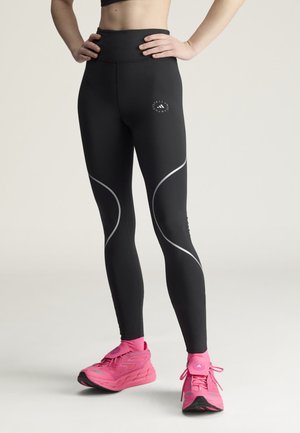 Kvinde iført sorte højtaljede leggings med refleksdetaljer og lyserøde sportssko, stående med hænderne på hoften.