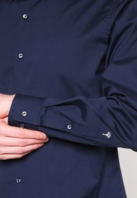 Granatowa koszula zapinana na guziki, uszyta z gładkiego materiału, charakteryzująca się eleganckim fasonem, metalowymi guzikami oraz subtelnym haftem na mankiecie.