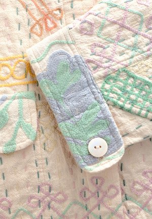 Beżowy materiał o fakturze z wielobarwną haftowaną dekoracją w pastelowych odcieniach zieleni, niebieskiego i żółtego. Zawiera guzik i ozdobną kieszeń.