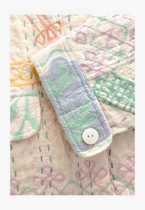 Beżowy materiał o fakturze z wielobarwną haftowaną dekoracją w pastelowych odcieniach zieleni, niebieskiego i żółtego. Zawiera guzik i ozdobną kieszeń.