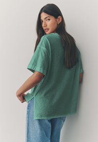 Zelené, nadmerné tričko s mäkkým materiálom, krátkymi rukávmi a voľným strihom, skombinované so svetlomodrými riflami.